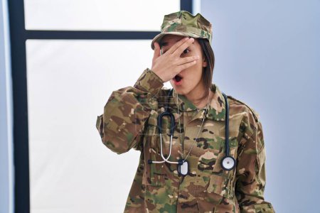 Téléchargez les photos : Jeune femme sud-asiatique portant un uniforme de camouflage de l'armée et stéthoscope regardant sous le choc couvrant le visage et les yeux avec la main, regardant à travers les doigts peur - en image libre de droit