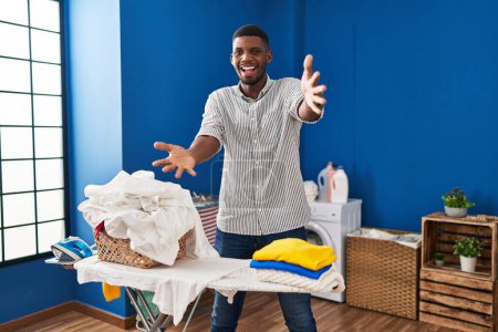 Téléchargez les photos : Homme afro-américain repassage vêtements à la maison en regardant la caméra souriant à bras ouverts pour étreindre. expression joyeuse embrassant le bonheur. - en image libre de droit