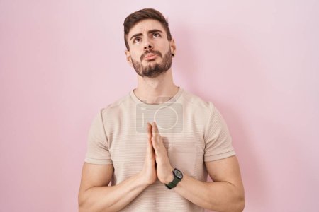 Téléchargez les photos : Hispanic man with beard standing over pink background begging and praying with hands together with hope expression on face very emotional and worried. begging. - en image libre de droit