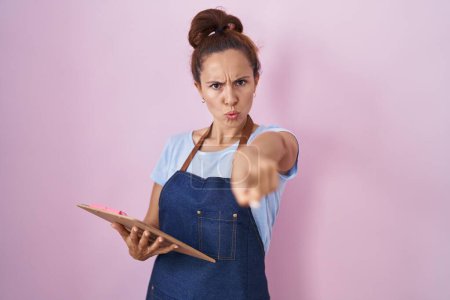 Photo for Brunette woman wearing professional waitress apron holding clipboard pointing with finger to the camera and to you, confident gesture looking serious - Royalty Free Image