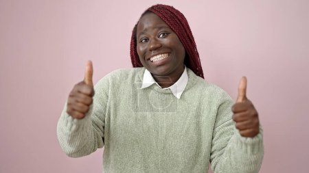 Téléchargez les photos : Femme africaine aux cheveux tressés souriant avec les pouces vers le haut sur fond rose isolé - en image libre de droit