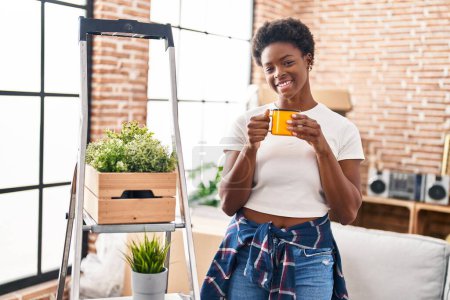 Téléchargez les photos : Femme afro-américaine souriante boire du café avec confiance à la nouvelle maison - en image libre de droit