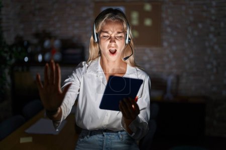 Photo for Young blonde woman working at the office at night doing stop gesture with hands palms, angry and frustration expression - Royalty Free Image