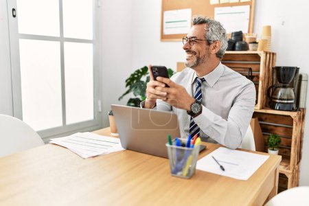 Téléchargez les photos : Homme d'affaires aux cheveux gris d'âge moyen utilisant un ordinateur portable et un smartphone au bureau - en image libre de droit