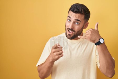 Photo for Handsome hispanic man standing over yellow background smiling doing talking on the telephone gesture and pointing to you. call me. - Royalty Free Image