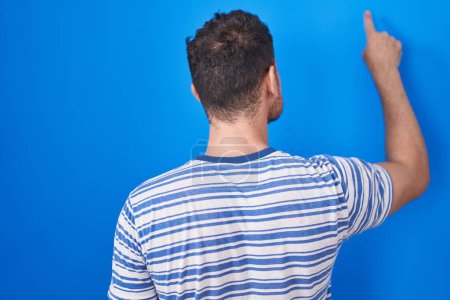 Foto de Joven hombre hispano de pie sobre fondo azul posando hacia atrás apuntando hacia adelante con la mano del dedo - Imagen libre de derechos