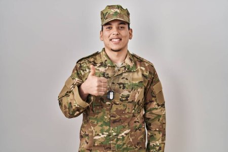 Téléchargez les photos : Jeune homme arabe portant un uniforme de camouflage de l'armée faisant des pouces heureux geste avec la main. approuvant l'expression regardant la caméra montrant le succès. - en image libre de droit