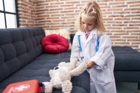 Photo for Adorable blonde girl playing doctor with teddy bear at home - Royalty Free Image