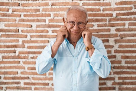 Photo for Senior man with grey hair standing over bricks wall covering ears with fingers with annoyed expression for the noise of loud music. deaf concept. - Royalty Free Image