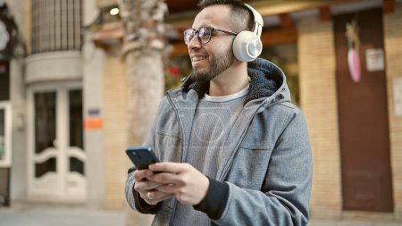 Téléchargez les photos : Hispanique homme souriant confiant écouter de la musique en utilisant smartphone à la rue - en image libre de droit