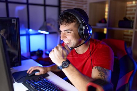 Téléchargez les photos : Jeune homme hispanique streamer jouer à un jeu vidéo avec l'expression gagnante à la salle de jeux - en image libre de droit
