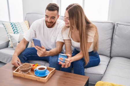 Foto de Hombre y mujer pareja desayunando usando smartphone en casa - Imagen libre de derechos
