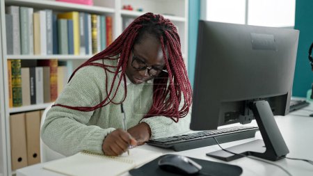 Téléchargez les photos : Femme africaine avec étudiant cheveux tressés en utilisant l'écriture informatique sur ordinateur portable à la bibliothèque universitaire - en image libre de droit