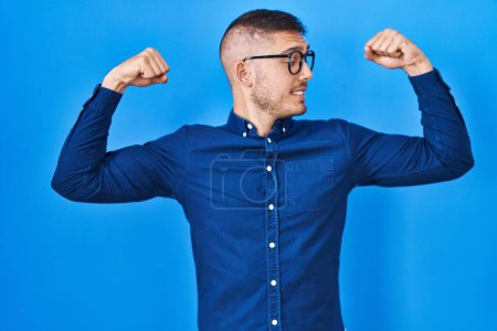 Foto de Joven hombre hispano con gafas sobre fondo azul mostrando los músculos de los brazos sonriendo orgulloso. concepto de fitness. - Imagen libre de derechos
