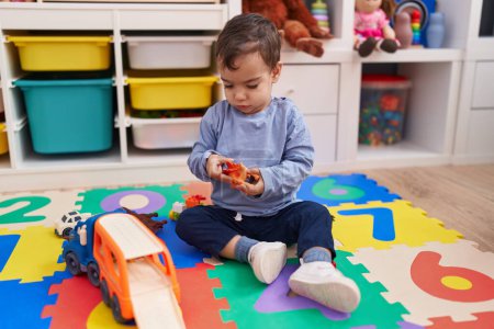 Téléchargez les photos : Adorable garçon hispanique jouant avec voiture et jouet de dinosaure assis sur le sol à la maternelle - en image libre de droit
