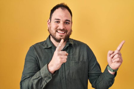 Photo for Plus size hispanic man with beard standing over yellow background smiling and looking at the camera pointing with two hands and fingers to the side. - Royalty Free Image
