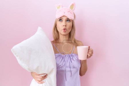 Foto de Mujer joven caucásica con pijama abrazando almohada y tomando café haciendo cara de pescado con boca y ojos entrecerrados, loca y cómica. - Imagen libre de derechos