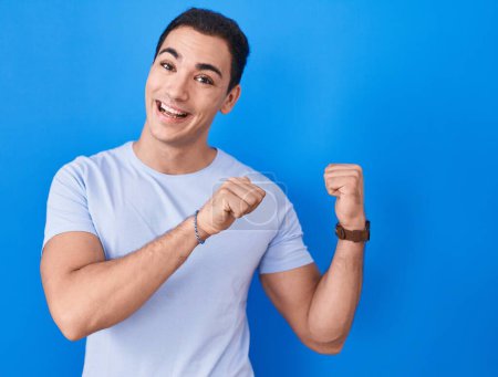 Foto de Young hispanic man standing over blue background pointing to the back behind with hand and thumbs up, smiling confident - Imagen libre de derechos