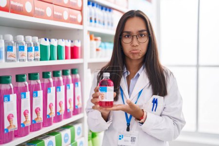 Foto de Mujer hispana joven que trabaja en farmacia sosteniendo jarabe deprimido y preocupada por la angustia, llorando enojada y asustada. expresión triste. - Imagen libre de derechos