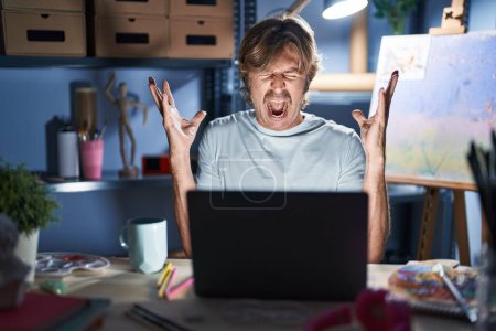 Photo for Middle age man sitting at art studio with laptop at night celebrating mad and crazy for success with arms raised and closed eyes screaming excited. winner concept - Royalty Free Image