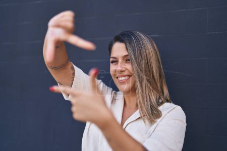 Foto de Joven mujer hispana sonriendo confiada haciendo gesto fotográfico con las manos de pie junto a la pared - Imagen libre de derechos