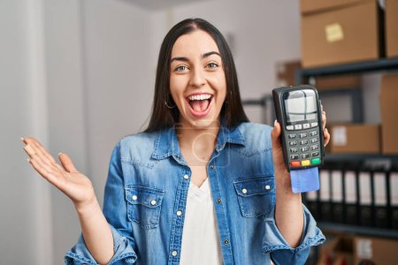 Téléchargez les photos : Femme hispanique travaillant au commerce électronique de petite entreprise tenant la carte de crédit et dataphone célébrant la victoire avec sourire heureux et expression gagnante avec les mains levées - en image libre de droit