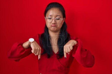 Foto de Mujer joven asiática de pie sobre fondo rojo apuntando hacia abajo mirando triste y molesto, indicando la dirección con los dedos, infeliz y deprimido. - Imagen libre de derechos