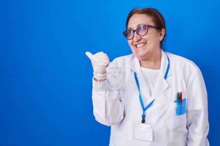 Foto de Mujer mayor con gafas vistiendo uniforme científico sonriendo con la cara feliz mirando y apuntando hacia un lado con el pulgar hacia arriba. - Imagen libre de derechos
