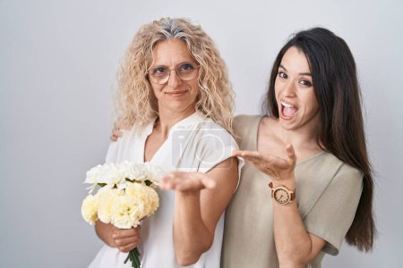 Téléchargez les photos : Mère et fille tenant bouquet de fleurs blanches pointant de côté avec les mains ouvertes paumes montrant l'espace de copie, présentant la publicité souriant heureux excité - en image libre de droit