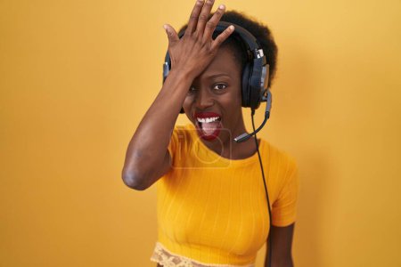Téléchargez les photos : Femme africaine avec les cheveux bouclés debout sur fond jaune portant des écouteurs surpris avec la main sur la tête pour erreur, rappelez-vous erreur. oublié, mauvais concept de mémoire. - en image libre de droit