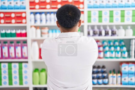 Téléchargez les photos : Jeune homme chinois travaillant à la pharmacie drugstore debout vers l'arrière regardant loin avec les bras croisés - en image libre de droit