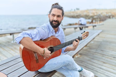 Photo for Young hispanic man musician playing classical guitar sitting on bench at seaside - Royalty Free Image