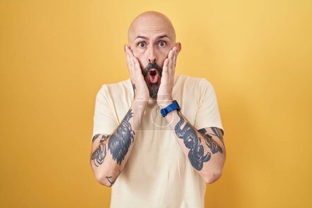 Foto de Hombre hispano con tatuajes de pie sobre fondo amarillo asustado y sorprendido, sorpresa y expresión asombrada con las manos en la cara - Imagen libre de derechos