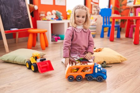 Photo for Adorable blonde girl playing with truck toy and dinosaur sitting on floor at kindergarten - Royalty Free Image