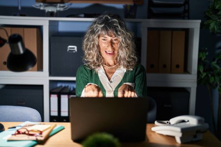 Photo for Middle age woman working at night using computer laptop pointing to you and the camera with fingers, smiling positive and cheerful - Royalty Free Image