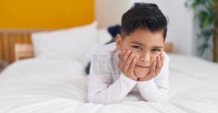 Téléchargez les photos : Adorable garçon hispanique couché sur le lit avec une expression sérieuse dans la chambre - en image libre de droit
