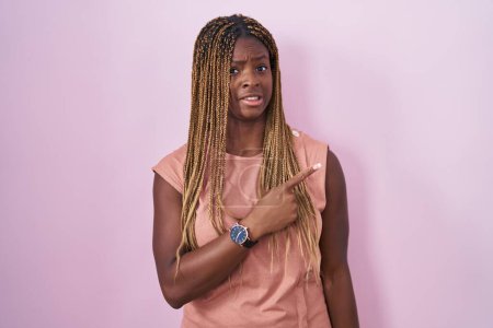 Téléchargez les photos : Femme afro-américaine avec des cheveux tressés debout sur fond rose pointant de côté inquiet et nerveux avec index, expression préoccupée et surprise - en image libre de droit