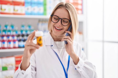 Téléchargez les photos : Jeune femme blonde pharmacien tenant bouteille pilules parler sur smartphone à la pharmacie - en image libre de droit