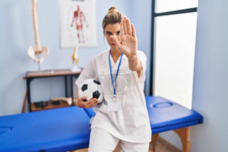 Téléchargez les photos : Jeune femme travaillant à la clinique de thérapie de football avec la main ouverte faisant stop signe avec expression sérieuse et confiante, geste de défense - en image libre de droit