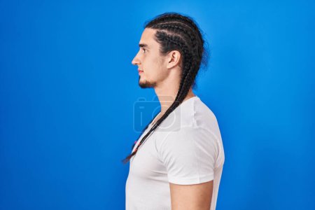 Téléchargez les photos : Homme hispanique aux cheveux longs debout sur fond bleu regardant vers le côté, pose de profil relax avec visage naturel avec sourire confiant. - en image libre de droit
