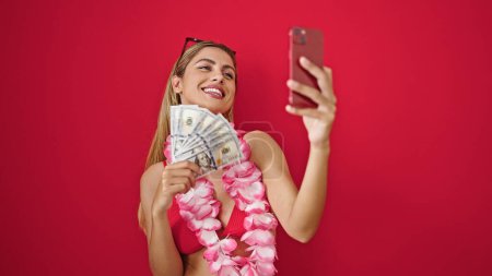 Foto de Joven mujer rubia con bikini sosteniendo dólares teniendo videollamada sobre fondo rojo aislado - Imagen libre de derechos