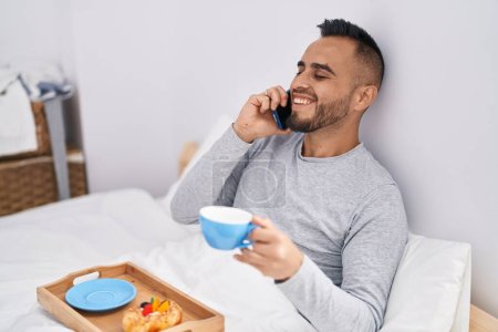 Téléchargez les photos : Jeune homme hispanique parlant sur smartphone prendre le petit déjeuner dans la chambre - en image libre de droit