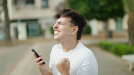 Photo for Non binary man using smartphone with winner expression at park - Royalty Free Image