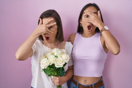 Foto de Madre e hija hispanas sosteniendo ramo de flores blancas asomándose en shock cubriendo la cara y los ojos con la mano, mirando a través de los dedos con expresión avergonzada. - Imagen libre de derechos