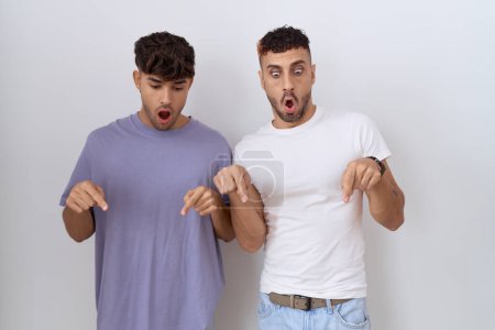 Téléchargez les photos : Couple gay homosexuel debout sur fond blanc pointant vers le bas avec les doigts montrant la publicité, visage surpris et bouche ouverte - en image libre de droit