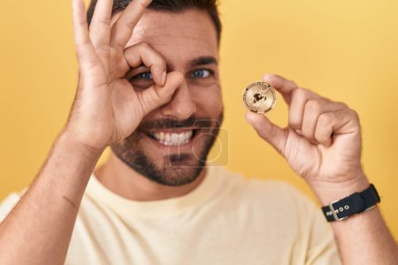 Foto de Hombre hispano guapo sosteniendo uniswap moneda criptomoneda sonriendo feliz haciendo signo ok con la mano en el ojo mirando a través de los dedos - Imagen libre de derechos