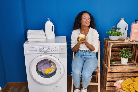 Photo for Young latin woman drinking coffee waiting for washing machine at laundru room - Royalty Free Image