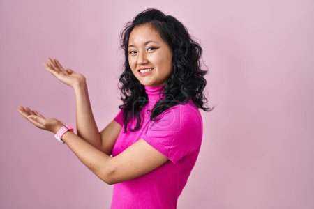 Foto de Joven asiática mujer de pie sobre rosa fondo invitando a entrar sonriendo natural con la mano abierta - Imagen libre de derechos