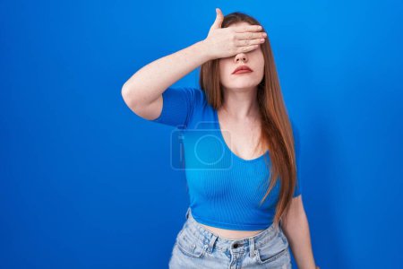 Photo for Redhead woman standing over blue background covering eyes with hand, looking serious and sad. sightless, hiding and rejection concept - Royalty Free Image