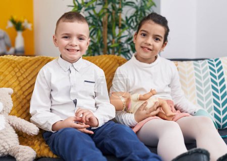 Téléchargez les photos : Adorable garçon et fille assis sur le canapé tenant bébé poupée à la maison - en image libre de droit